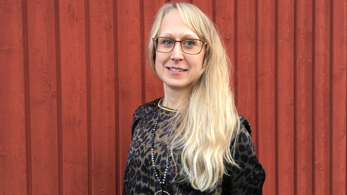 Anna Lindgren har ljust långt hår och står med en stor röd ladugårdsvägg bakom sig