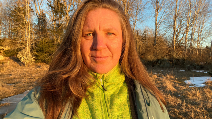 Elisabeth Bölenius har grön tröja och jacka på sig och hon står utomhus i starkt solljus