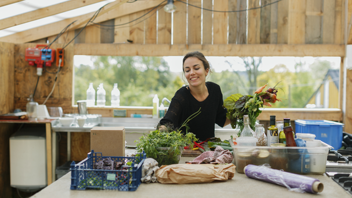 Kvinna i ett uthus står vid en bänk och plockar ihop grönsaker.
