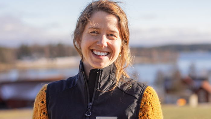 Lovisa Granberg har en orange tröja och en mörk väst på sig och står ute i naturen