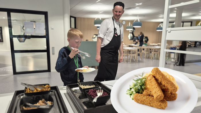 Ett barn tar mat från en buffé med en kock ståendes bredvid. Infällt i bild är en tallrik med fiskpinnar.