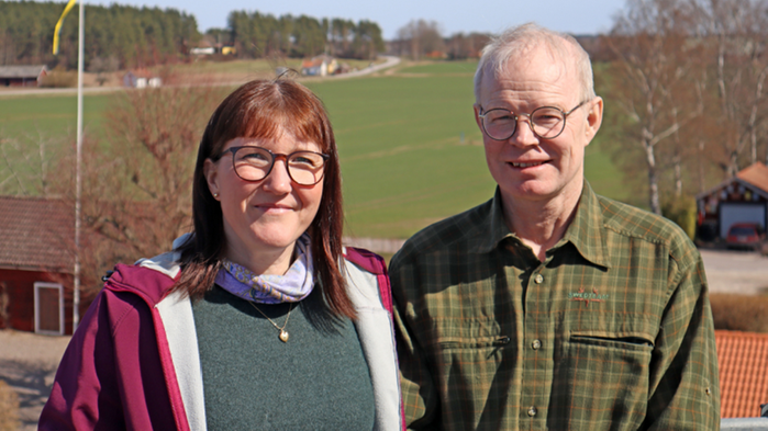 Karolina Muhrman och Per Frankelius står vid en röd stuga med ett fält i bakgrunden